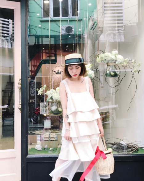 Cô chủ shop điệu đà của cửa hàng thời trang REN biết rõ bản thân hợp nhất trong những chiếc đầm xòe dịu dàng. Chiếc đầm trắng xếp tầng đi với túi cói thắt dây lụa đỏ chính là điểm nhấn cho sự ngọt ngào của cô.