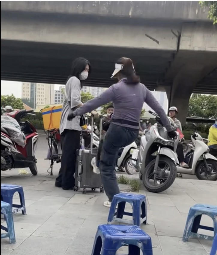 Hình ảnh người phụ nữ đá vào vali của cô gái đứng trên vỉa hè.