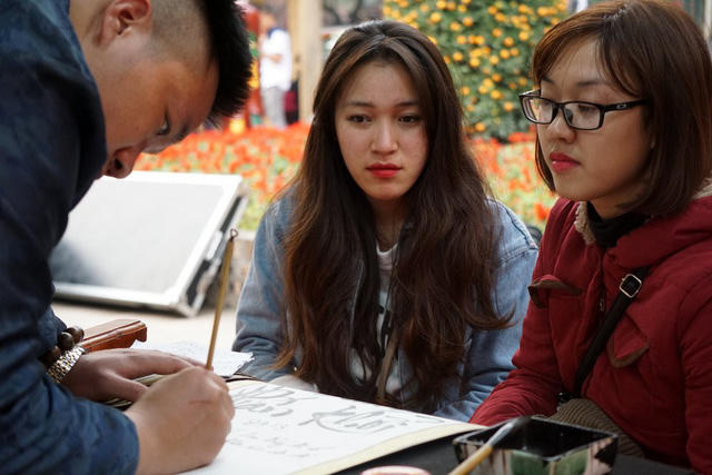 Hai bạn trẻ chăm chú theo dõi ông đồ viết thư pháp bằng chữ quốc ngữ.