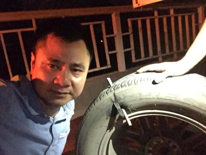 NSND Tự Long bên cạnh chiếc lốp xe ô tô bị thanh sắt đâm xuyên qua lốp.