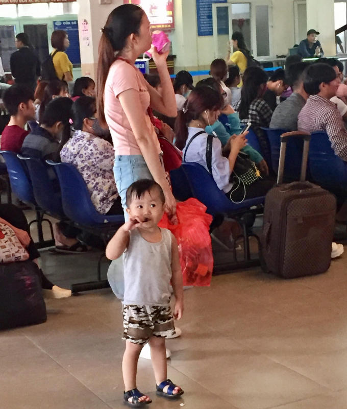 Hình ảnh bé trai ngơ ngác nhìn mọi người xung quang ở bến xe, vì ghế chờ hết nên em đứng cùng mẹ xem lịch trình đi của xe khách.