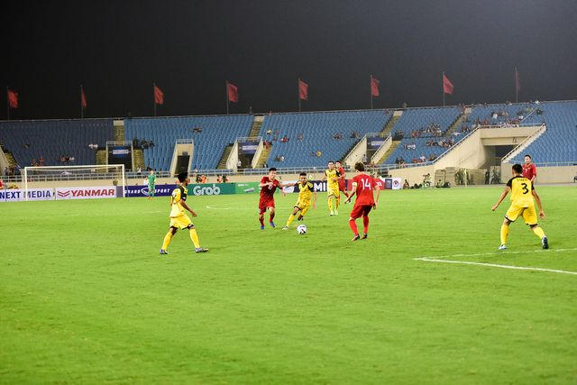 Ở góc máy này, chúng ta có thể thấy các cầu thủ đang thi đấu dưới sân, phía đằng xa trên khán đài có lác đác cổ động viên chủ yếu là nhiều ghế không có người ngồi.