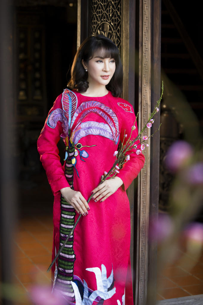 MC Thanh Mai chọn áo dài đỏ cho ngày đầu năm với hy vọng những điều tốt đẹp, may mắn sẽ đến. Trên nền chất liệu voan mỏng, họa tiết hoa lá bằng ren đắp nổi vừa tạo vẻ kín đáo, vừa tôn nét mềm mại, nữ tính cho người đẹp.