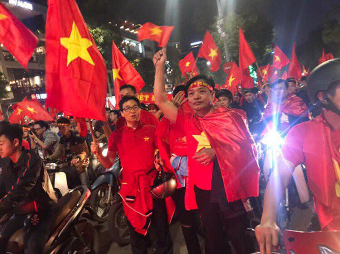 Phó Thủ tướng Vũ Đức Đam giản dị trong chiếc áo thun in ngôi sao vàng ở trái tim, mang theo lá cờ Tổ quốc chung vui với người hâm mộ bóng đá nước nhà.
