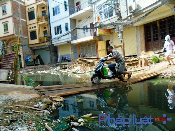 Người dân bắc các tấm ván lên mặt đường để vượt qua dòng nước đen ngòm.