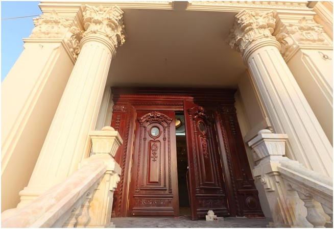 Cửa chính của biệt thự được làm bằng gỗ đổ, chạm khắc công phu. Hai bên có cột trụ sơn trắng và tay vịn bằng đá hoa cương đồng màu.