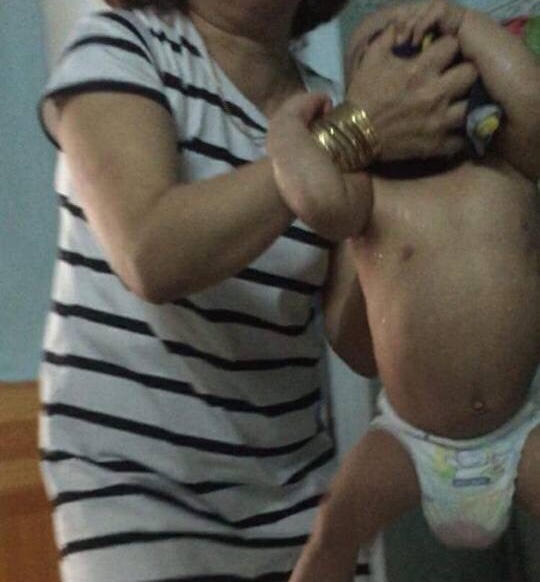 Bà Hồng dùng hai tay cầm vào đầu để nhấc bổng một bé trai ra khỏi cũi, được xác định là T.N.B.D. (12 tháng tuổi).