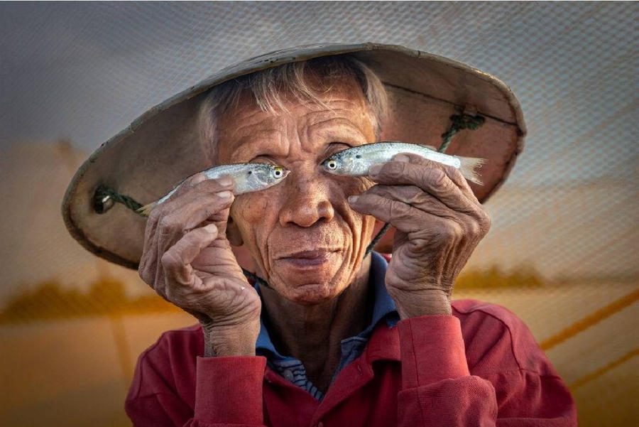 Tác phẩm "Mắt cá" (Fish Eyes) của Sophia Spurgin. "Bức ảnh ghi lại khoảnh khắc một ngư dân Hội An hóm hỉnh cầm hai con cá che trước mắt, phía sau là tấm lưới đánh cá thân thuộc. Tôi thực hiện tác phẩm này ngay trên thuyền khi buổi chụp hình sắp khép lại như một kỷ niệm vui vẻ đầy ngẫu hứng", tác giả xúc động nhớ lại. Khoảnh khắc xuất thần này đã giúp nhiếp ảnh gia Sophia Spurgin vượt qua hàng ngàn đối thủ để giành ngôi vị cao nhất cùng giải thưởng trị giá 3.000 bảng Anh.